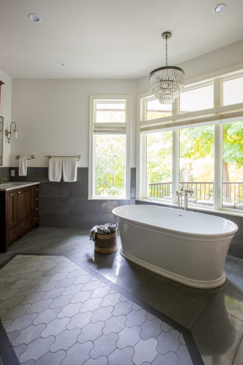 Expansive mullioned windows illuminate freestanding bathtub beneath crystal chandelier.