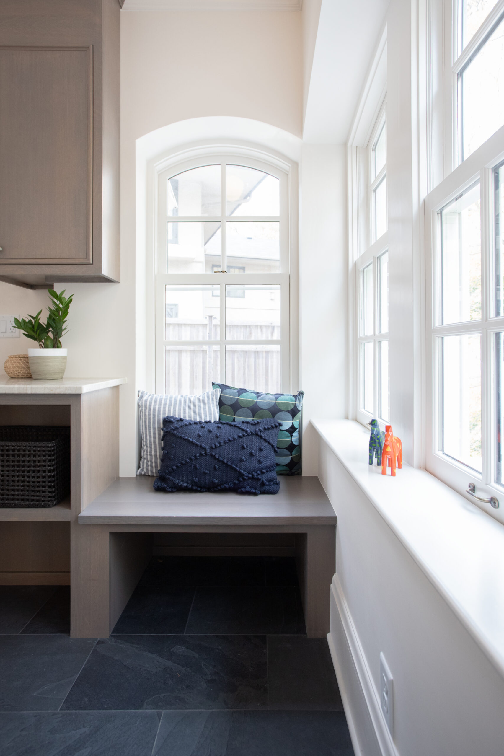 Arched-window mullioned glass floods oak-built-in benchseat, honed-slate flooring, textured-cushions.