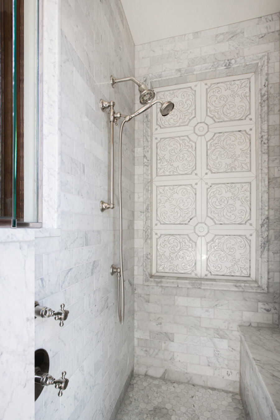 Expansive Carrara marble shower renovation features ornate panel, dual brushed nickel fixtures.
