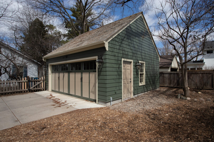 Renovated dark-green-sided garage features tan-paneled door, concrete drive, exterior lanterns.