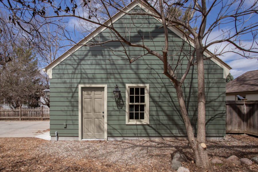 A detached green clapboard building features a beige paneled door, grid window, and wall sconce. Exterior home renovation.