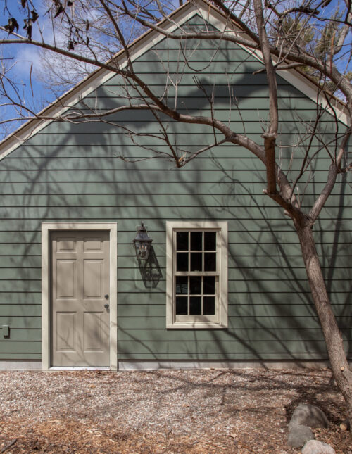 A detached green clapboard building features a beige paneled door, grid window, and wall sconce. Exterior home renovation.