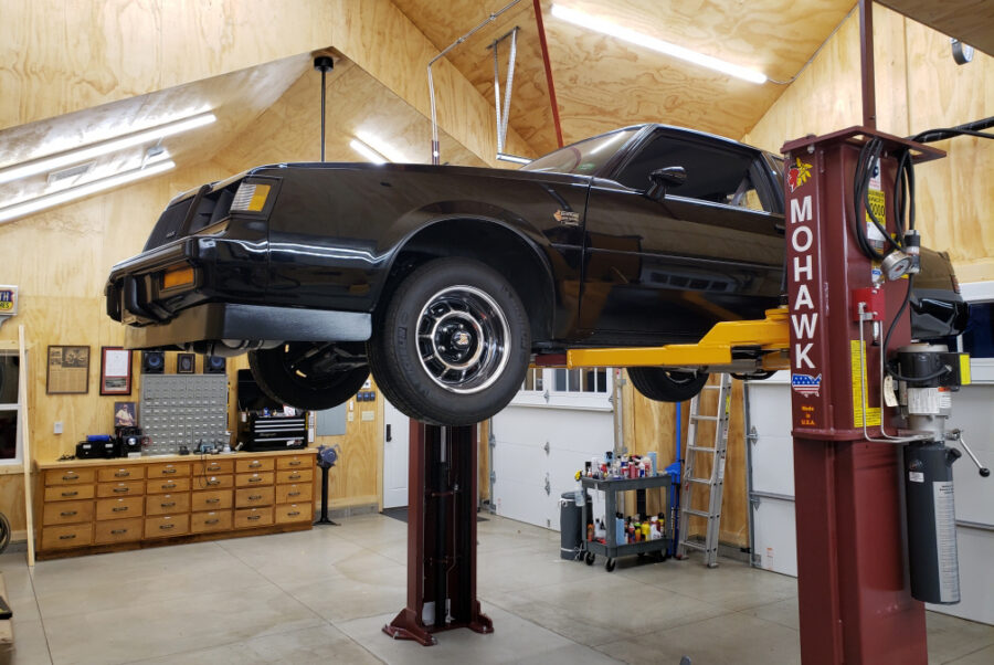 Plywood-paneled fluorescent-lit workshop features two-post lift hoisting gloss-black coupe
