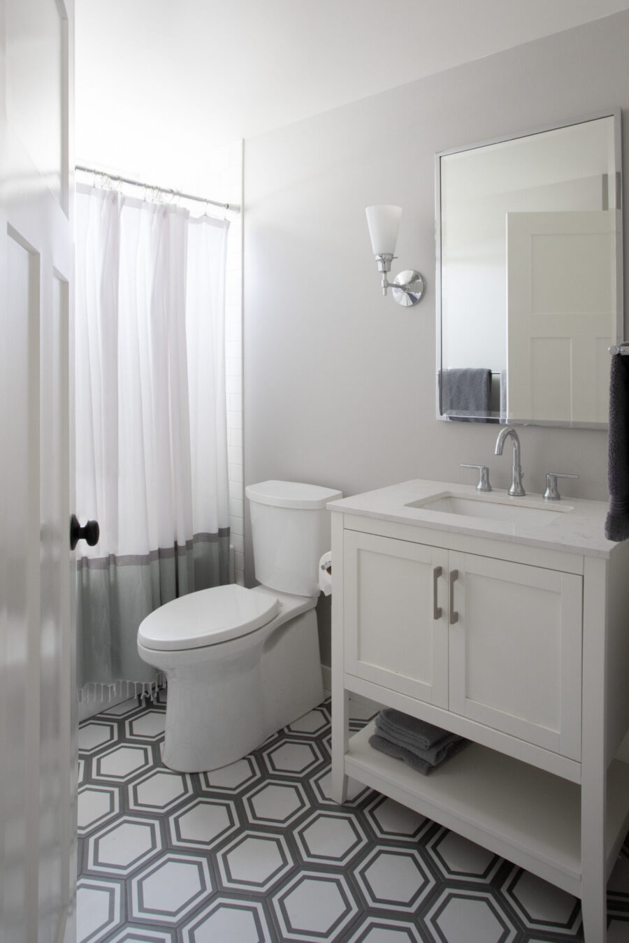 This bathroom remodel integrates hexagonal patterned floor tiles, white vanity, and a wall sconce.