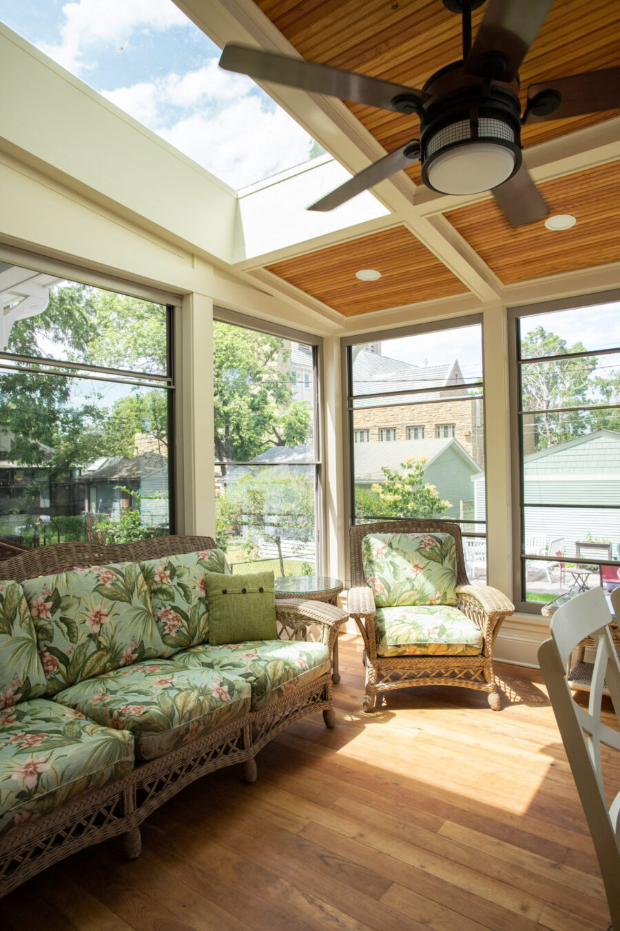 Bright Twin Cities sunroom addition features wicker seating, floral cushions, warm wood floors, and a skylit wood ceiling.