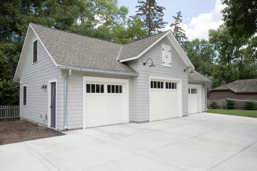 Newly renovated grey shingle garage boasts three white paneled doors, concrete drive.