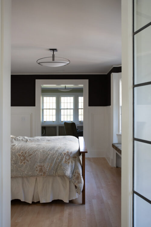 Circular-ceiling light hangs above solid-wood bedframe, paneled-wainscoting, hardwood-flooring