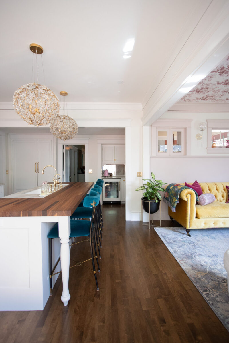 Bright open-concept kitchen remodel: wood island, teal stools, gold crystal lights, dark floors, and a yellow sofa.