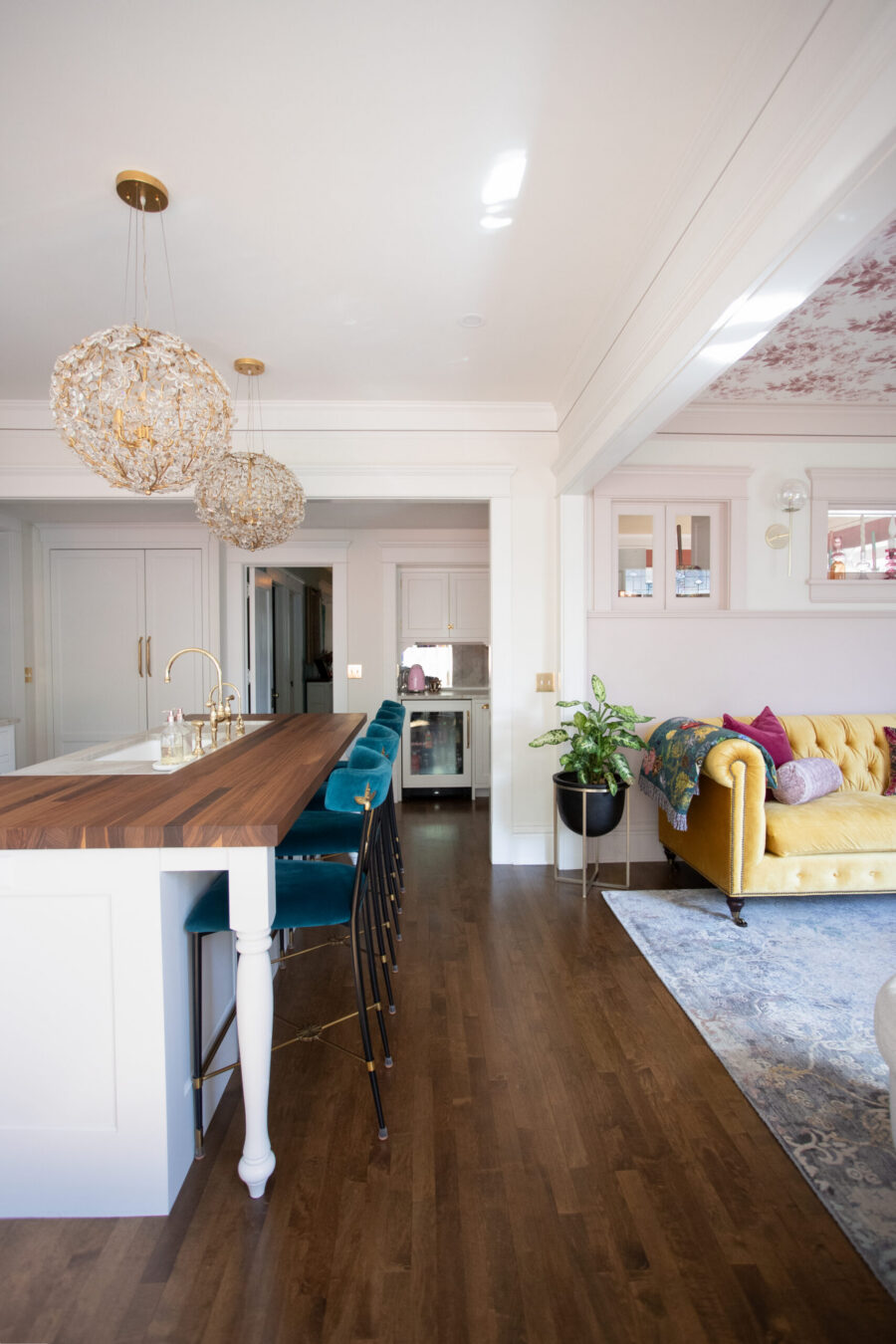 Bright open-concept kitchen remodel: wood island, teal stools, gold crystal lights, dark floors, and a yellow sofa.