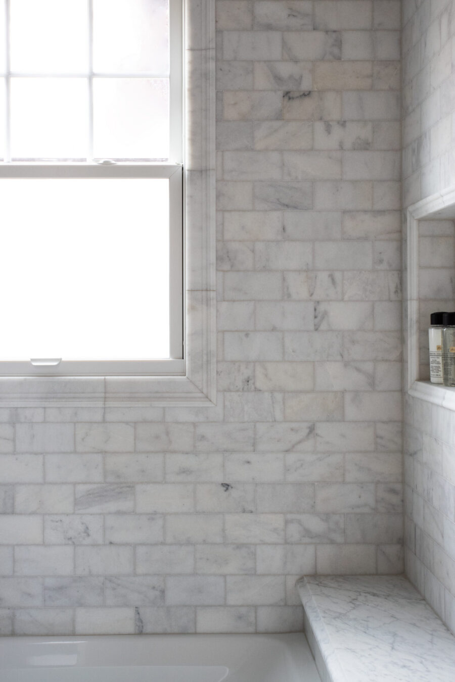 White marble bathroom renovation features bright window, recessed shelf, and shower-bench.