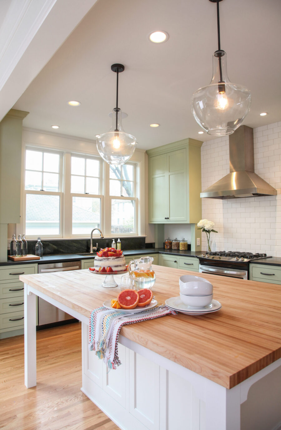 Remodeled kitchen boasts green cabinetry, wood island, subway tile, and elegant pendant lights.