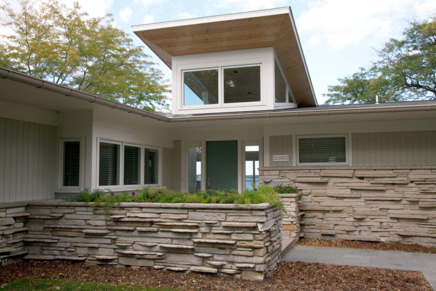 Modern design-build utilizes stacked stone, white siding, wood soffit, teal door, glass.