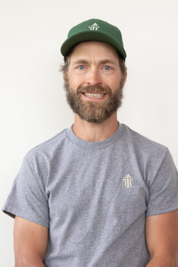 Smiling bearded professional, green-capped, grey-shirted, under even studio light.