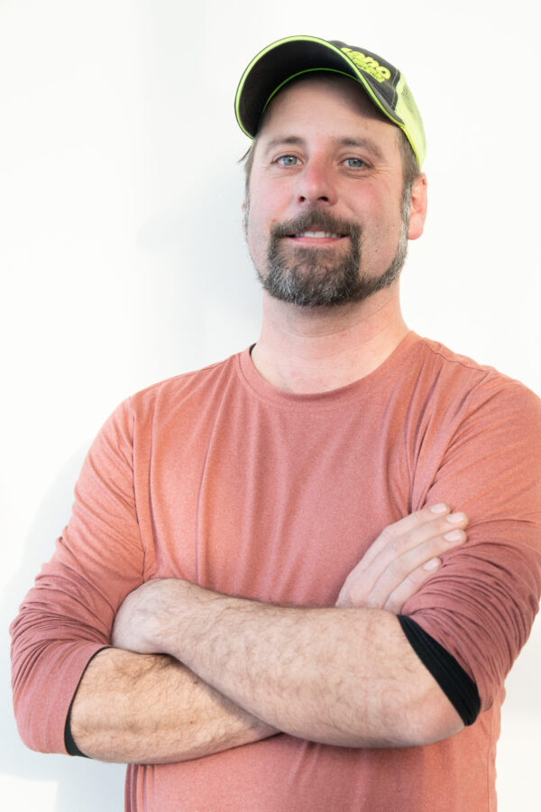 Smiling, bearded design-build professional, vibrant green-black cap, brick-red long-sleeve.
