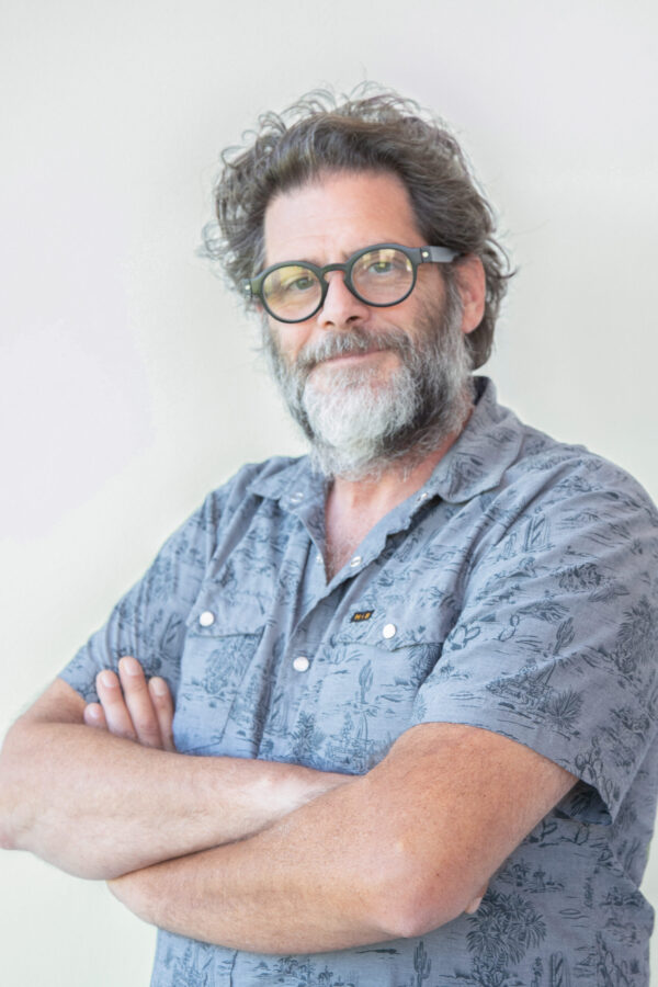 A grey-bearded man with glasses and a patterned shirt stands, crossing his arms on a light background.