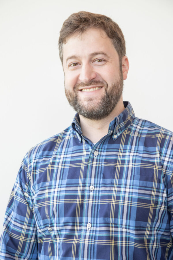 A smiling man with a beard and brown hair wears a blue plaid shirt. A bright white background is behind him.