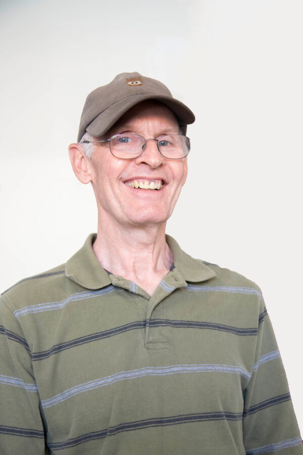 Man smiles, wearing glasses, a striped green polo shirt, and a brown baseball cap with an emblem.