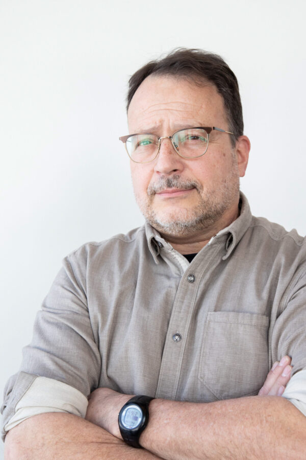 Professional man wears glasses, beard, tan collared shirt with rolled sleeves, arms crossed. Bright white interior lighting.