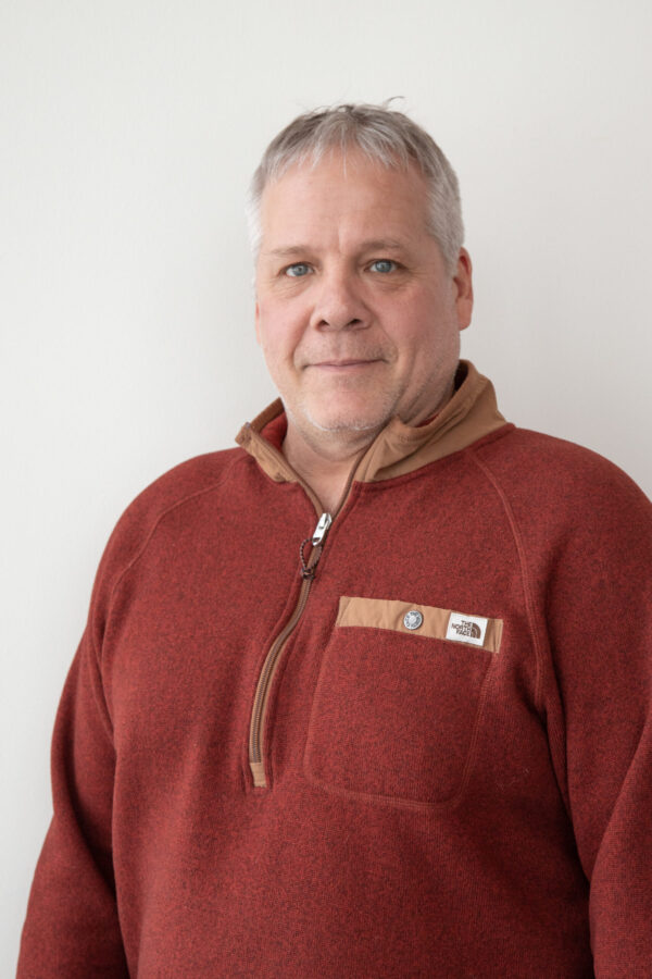 Gray-haired, blue-eyed man, reddish-brown quarter-zip fleece, bright studio-lighting background.