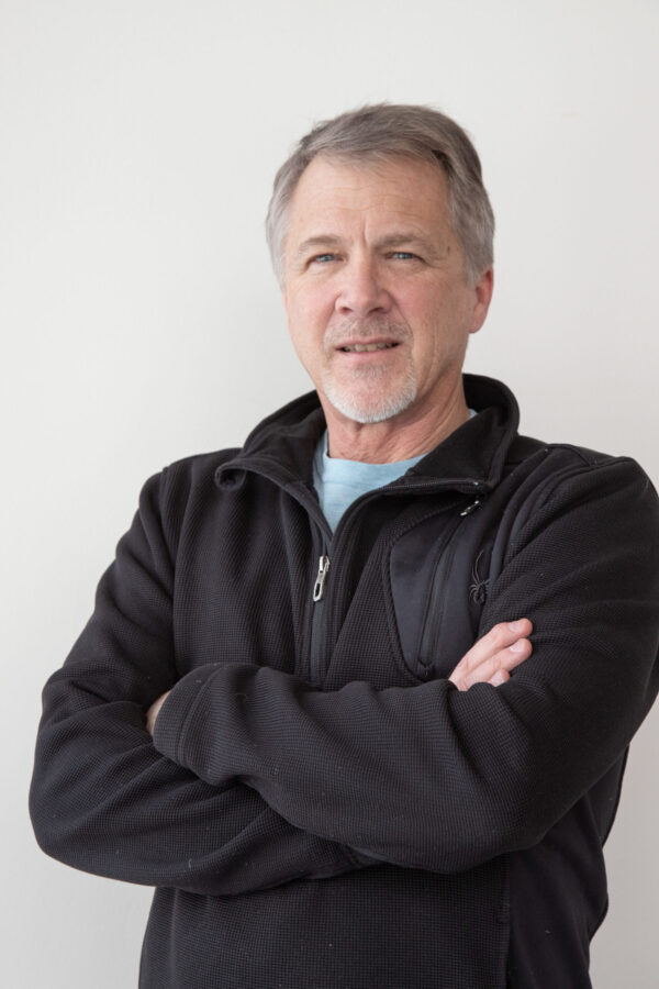 A man with grey hair and a goatee wears a black fleece jacket, arms confidently crossed.