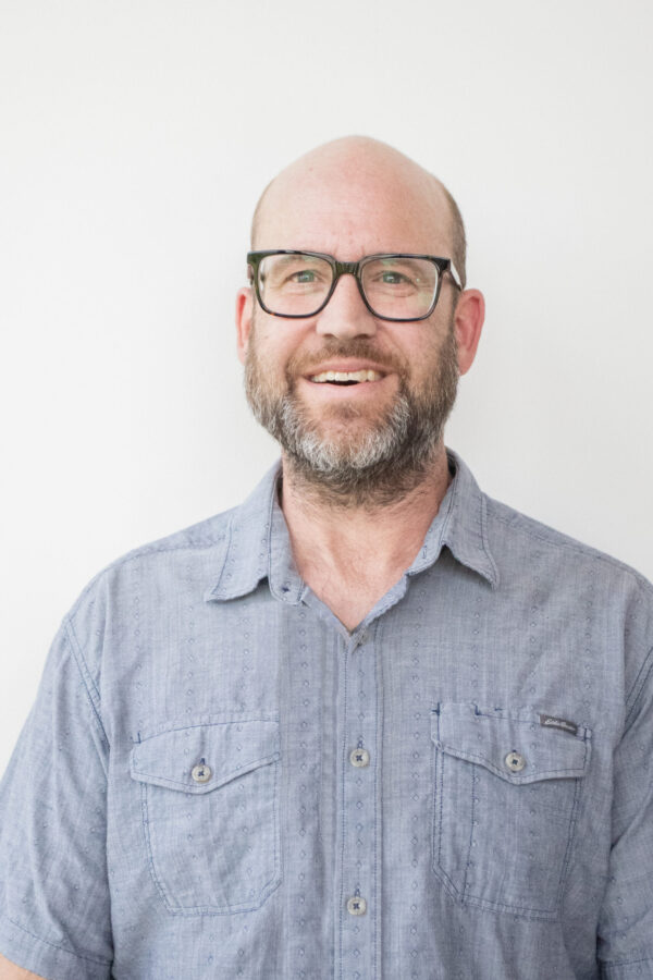 A smiling bald man with a short beard wears black-framed glasses and a light blue button-up shirt.