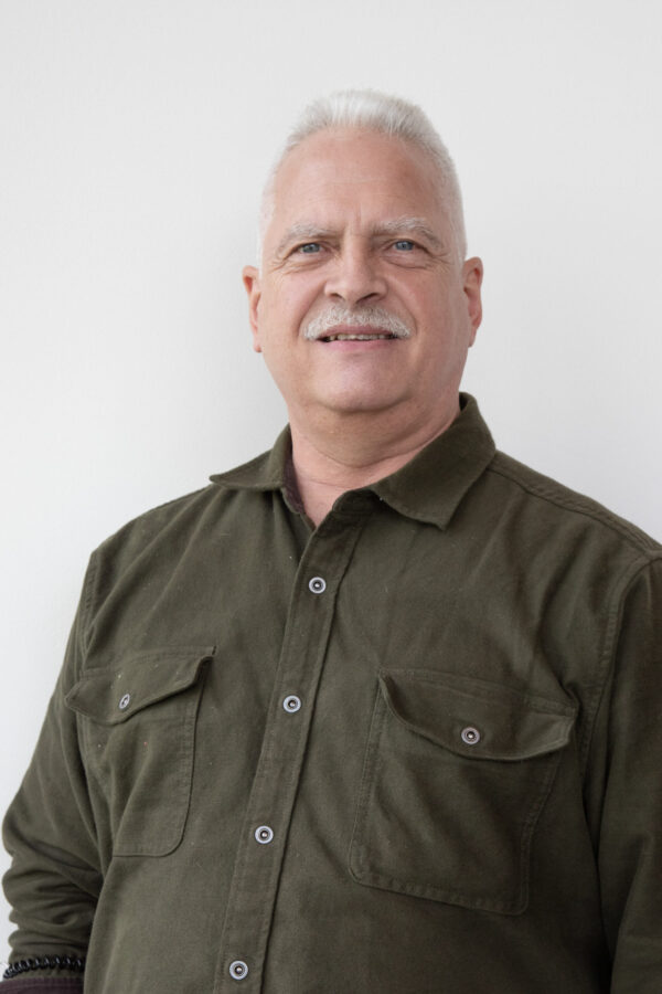 Smiling man with white hair and mustache wears a dark olive green collared shirt with two chest pockets. Subtle lighting highlights him against a plain white wall.