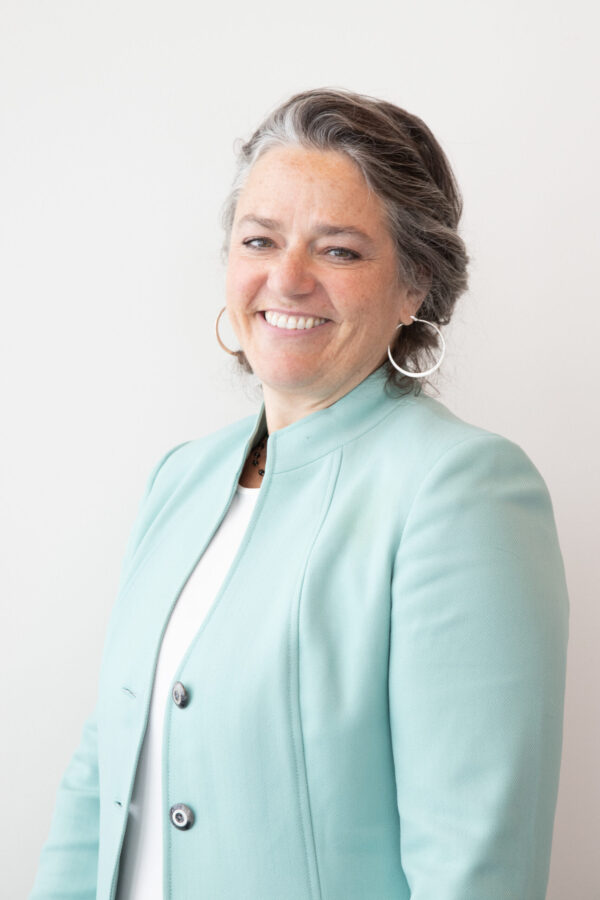 Smiling woman, gray hair, light teal blazer, white top, and large silver hoop earrings.