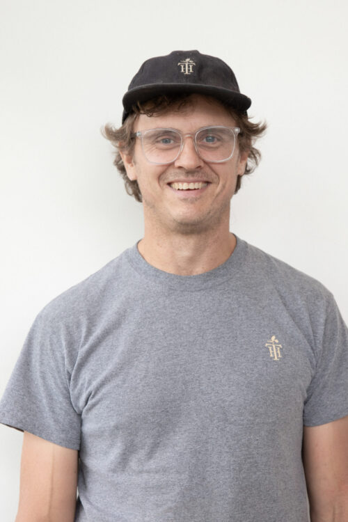Smiling man wears clear glasses, a dark baseball cap, and a gray t-shirt with a subtle chest design, against a plain white backdrop.