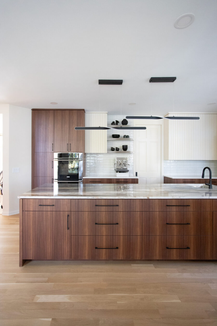 Kitchen remodel features rich walnut cabinetry, a large marble island, and sleek black pendants.