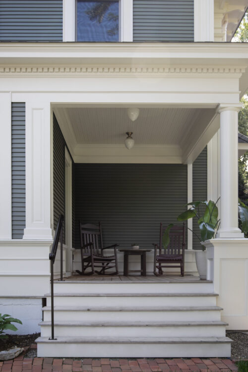 Classical-columned porch has beadboard tongue-and-groove ceiling, pendant-light fixtures, dark-wood-rockers
