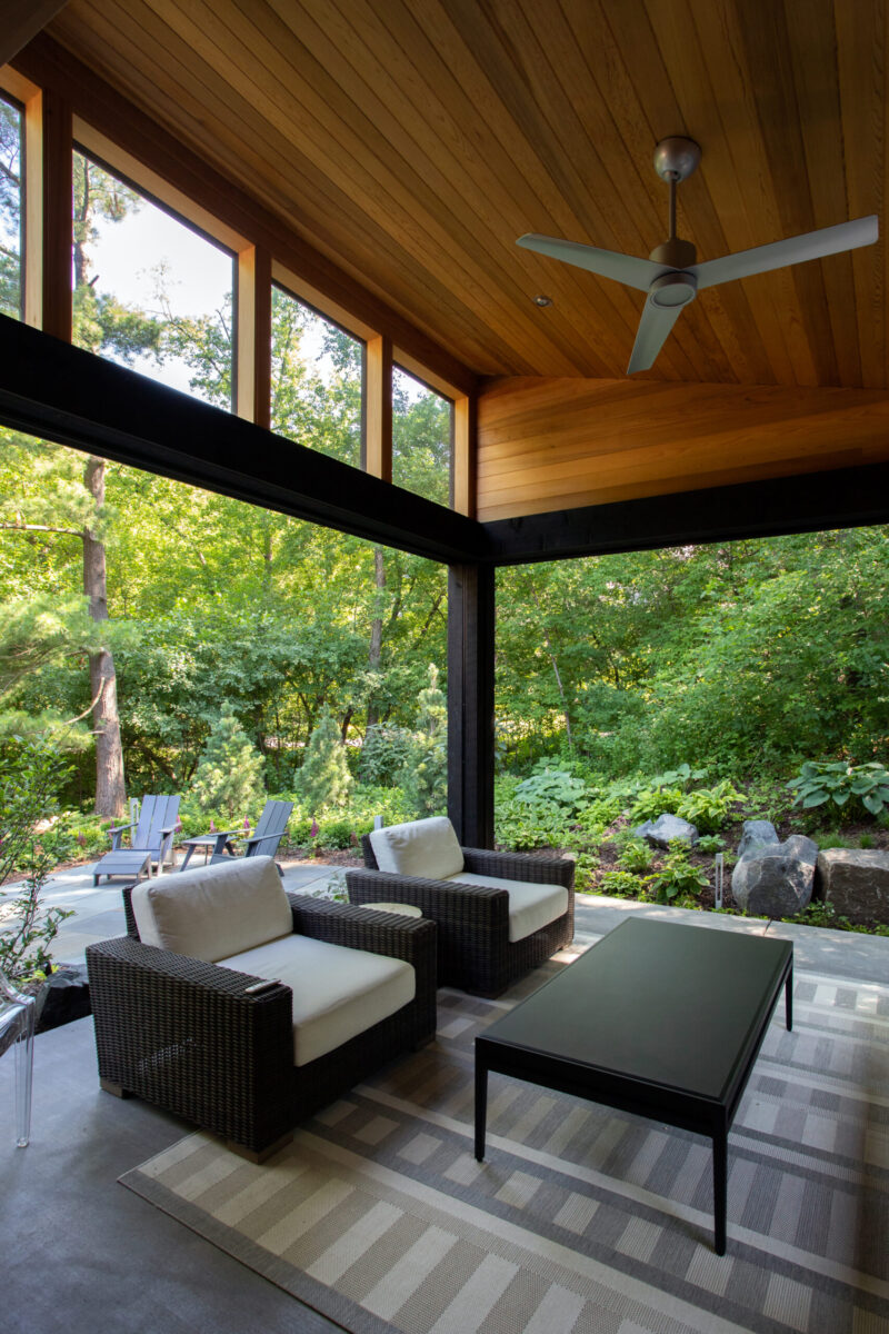 Modern patio has wood ceilings, dark beams, large windows. Outdoor seating on concrete views lush greenery.