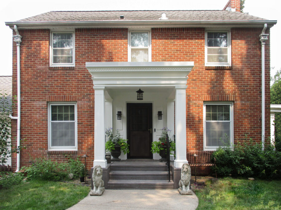 Stately red brick home exterior features prominent white portico, dark steps, lush landscaping.