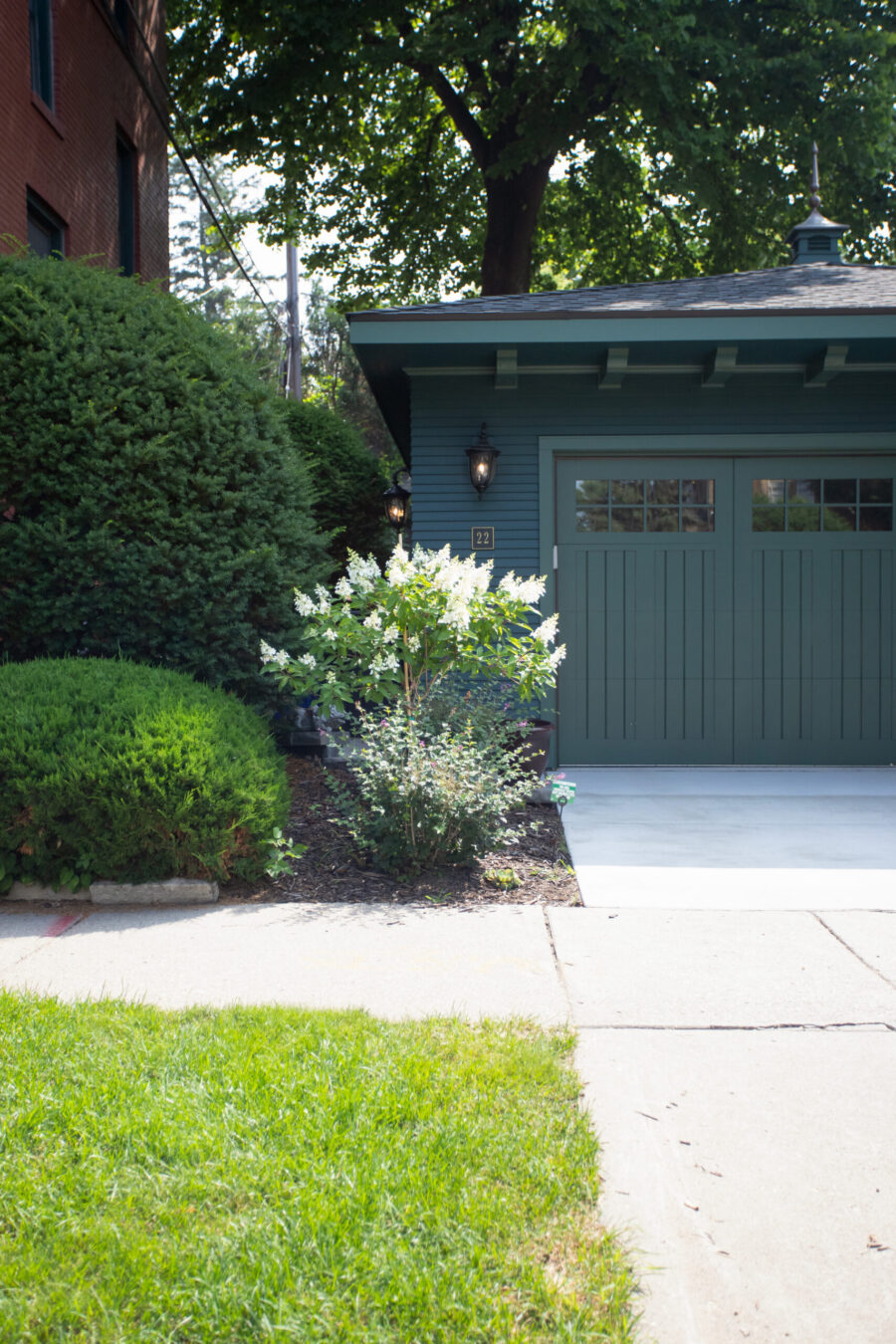 Green garage features two doors; bracketed eaves, dual wall lanterns, lush shrubs.