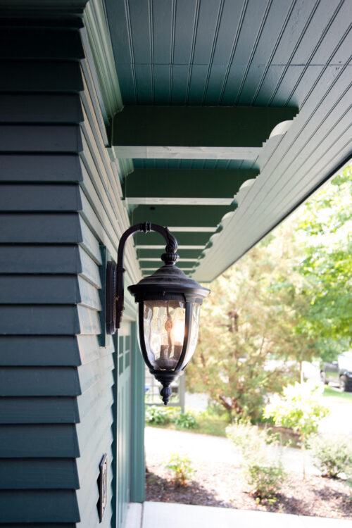 Dark teal horizontal siding, beadboard soffit, and a glowing wall lantern.