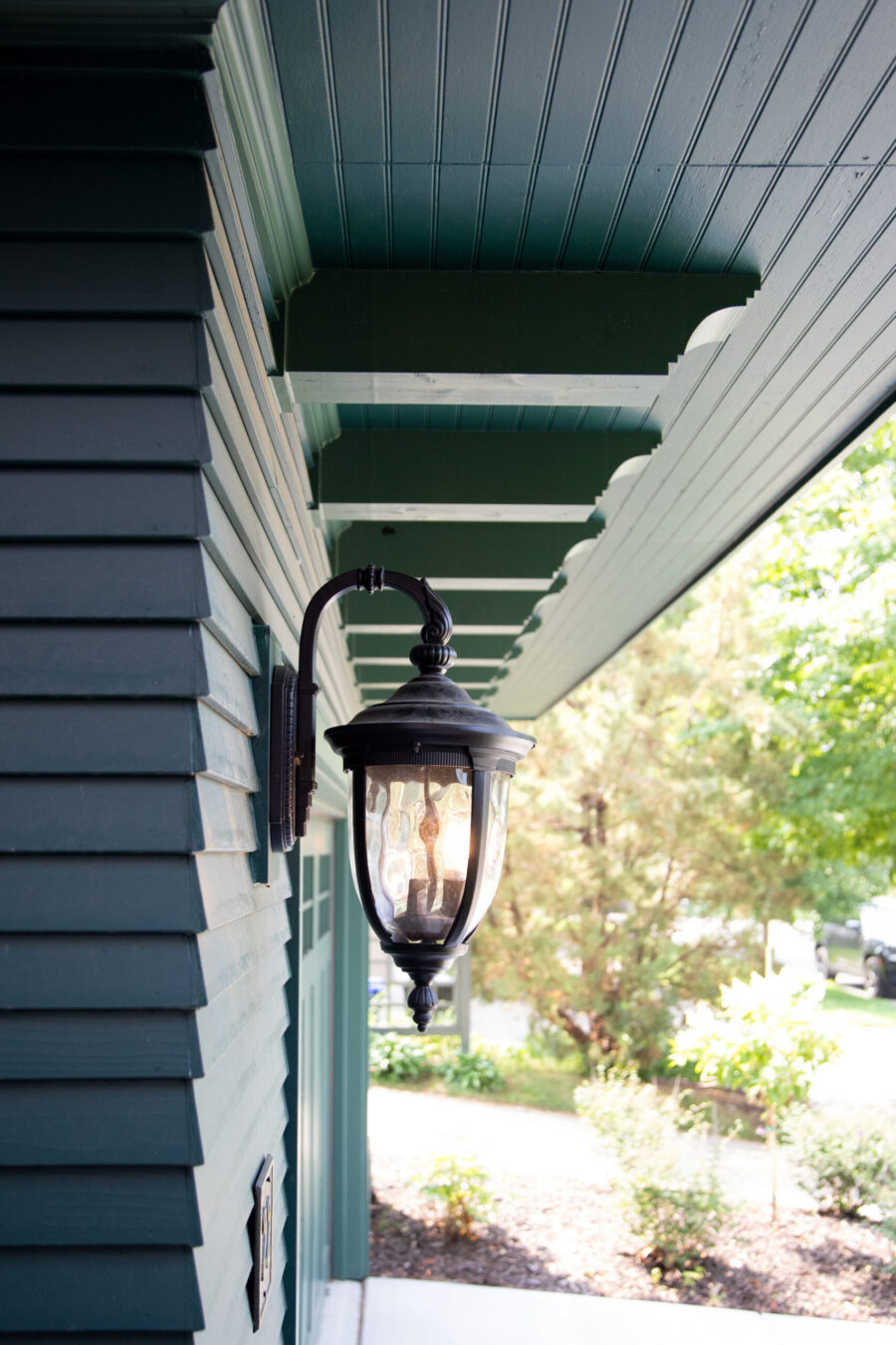 Dark teal horizontal siding, beadboard soffit, and a glowing wall lantern.