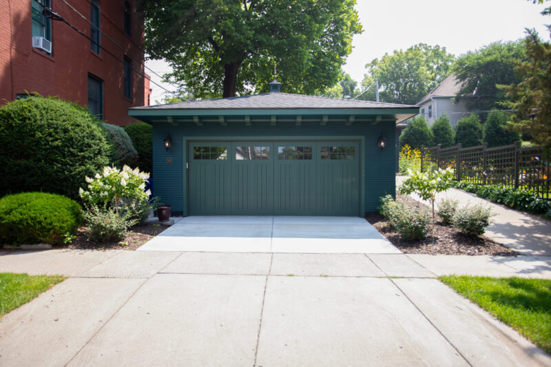 Green Craftsman garage has a light door, windows. Sconces illuminate. Driveway, landscaping. Exterior remodel.