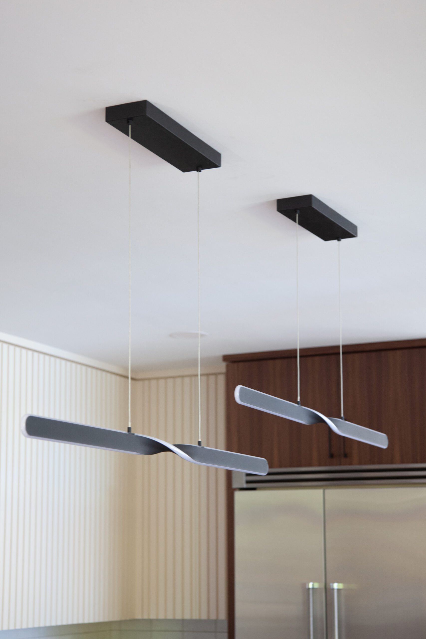 Two brushedstainlesswaveformpendantfixtures hang from matteblackrectangular ceilingplates above stainlessrefrigerator