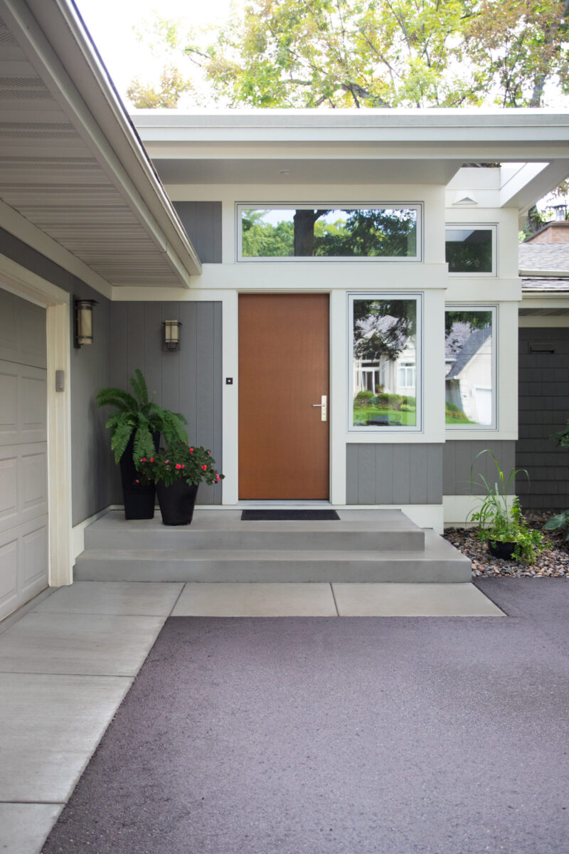 Renovated grey modern home displays brown door, concrete entry, outdoor lighting.
