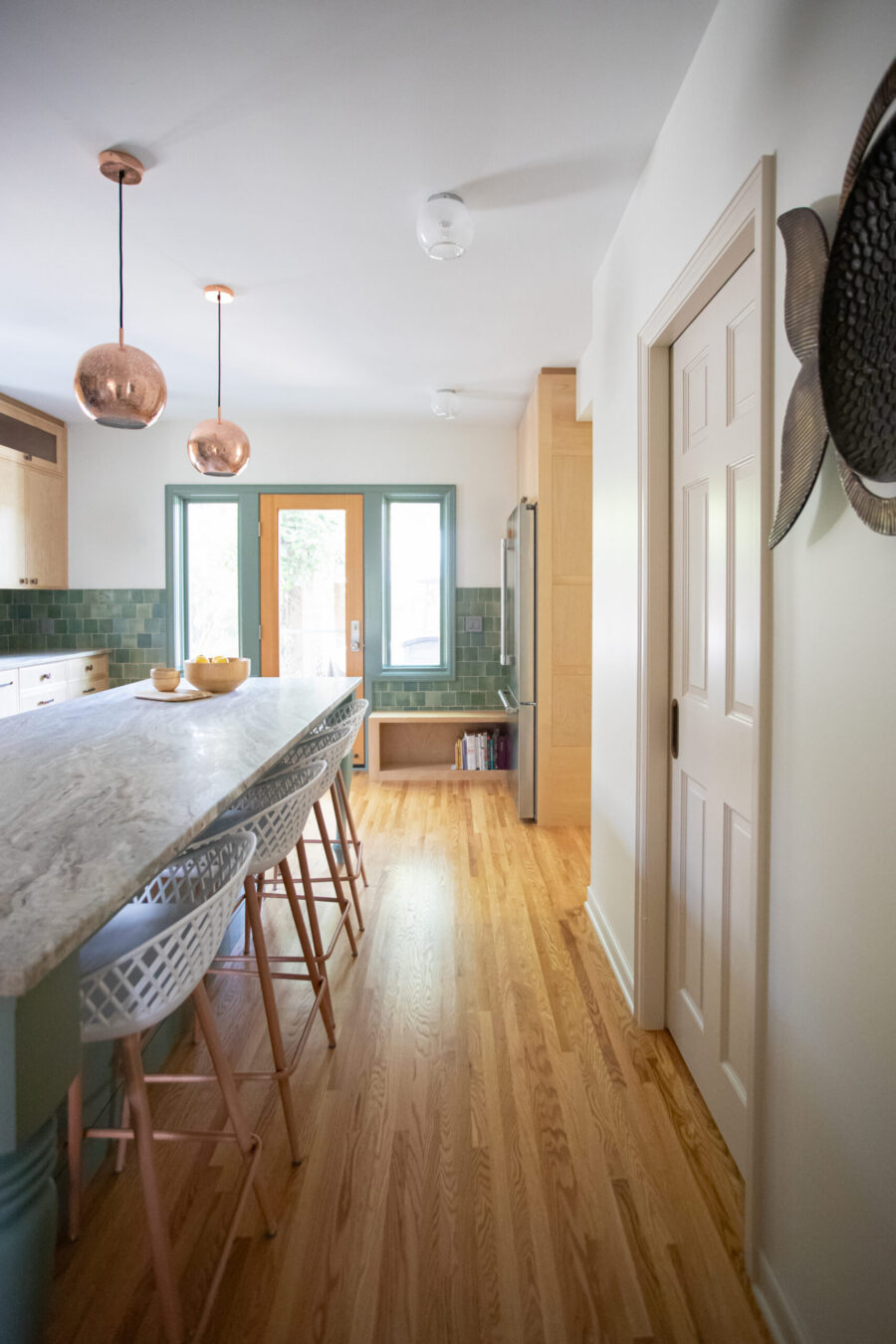 Renovated kitchen displays marble island, copper pendants, green backsplash, and light wood flooring.