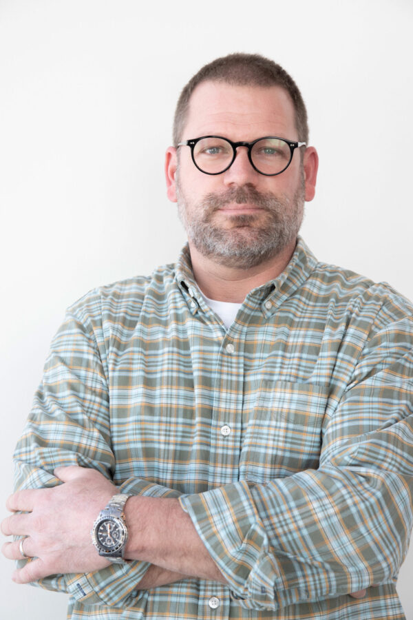 A bearded man wearing black-rimmed glasses and a green plaid shirt stands with crossed arms.