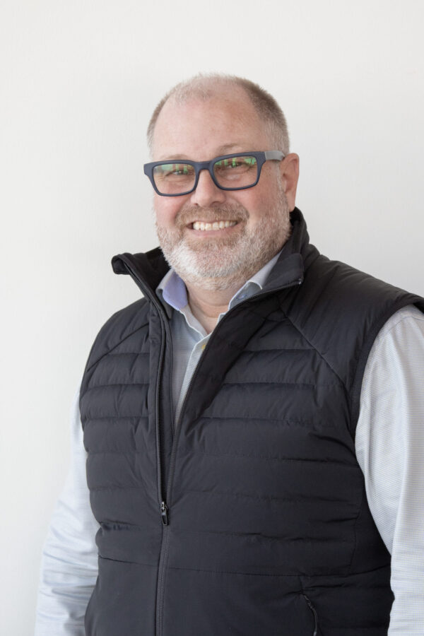 Bright, even lighting illuminates a smiling man's face, dark glasses, collared shirt, and black quilted puffer vest.
