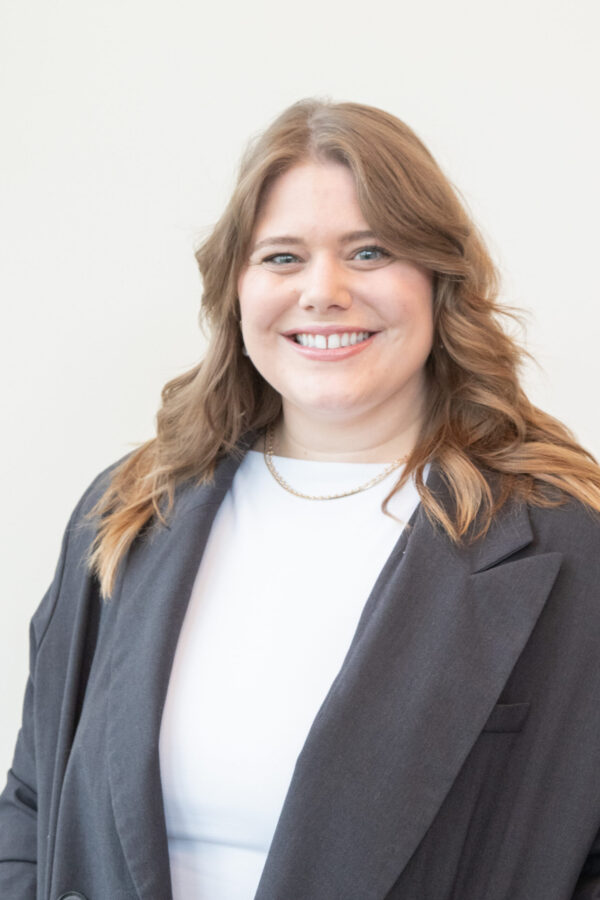 Smiling woman, long brown hair, dark gray blazer, white top, light wall.