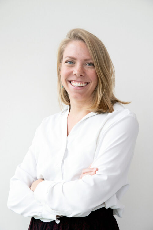 Smiling woman, blonde hair, white button-down shirt, arms crossed, bright white background.