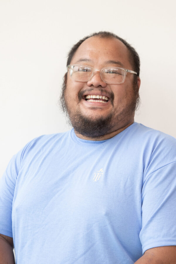 Cheerfully-grinning, bearded man wears spectacles, blue shirt against plain background.