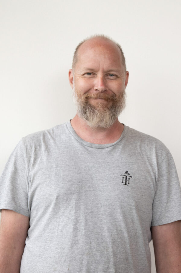 A man with a gray beard and short hair wears a plain gray t-shirt. Soft, even light illuminates his face and the light background.