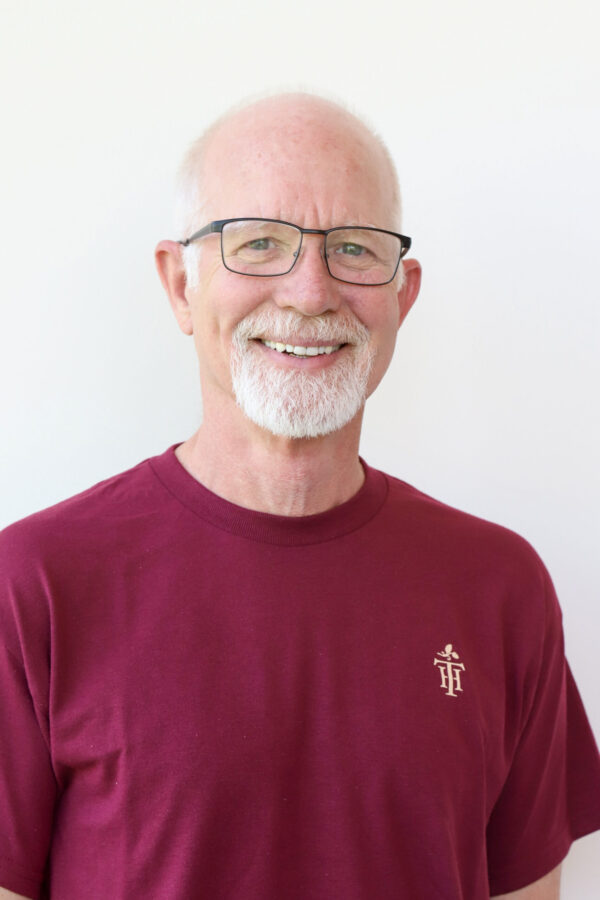 Smiling, bald man with a white goatee and black-framed glasses wears a maroon t-shirt on a plain white background.