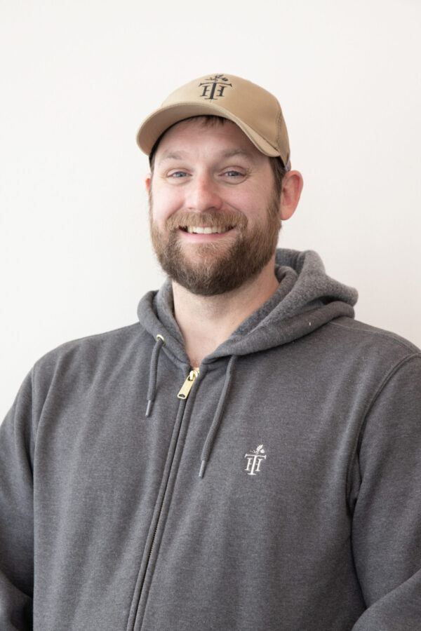 Smiling bearded man wears beige baseball cap and grey zippered hoodie. Plain white wall background.