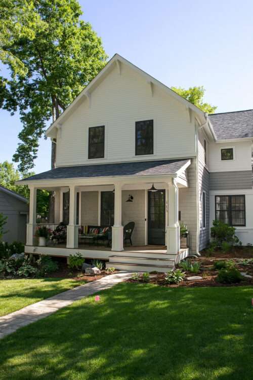 This white and grey modern home renovation features a wide front porch, dark windows, and vibrant landscaping.