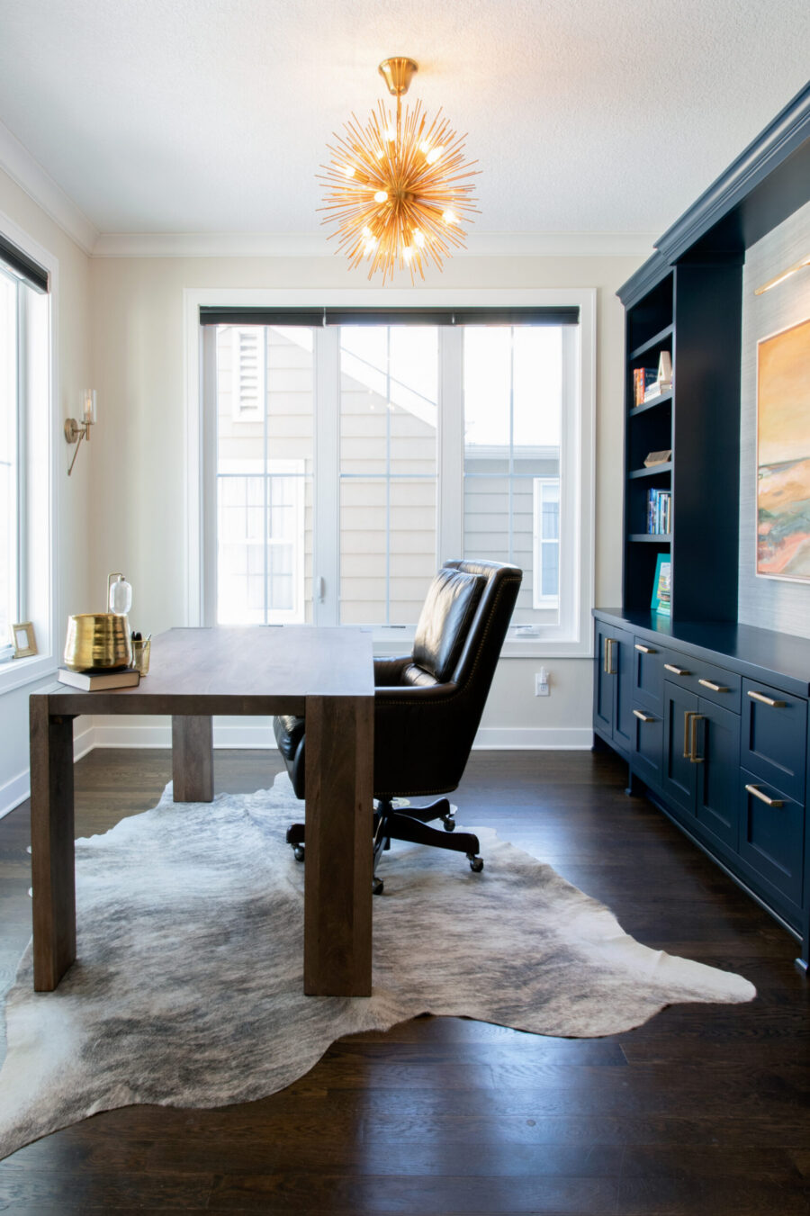 Twin Cities design build features dark wood desk, navy built-ins, gold chandelier, and a cowhide rug.