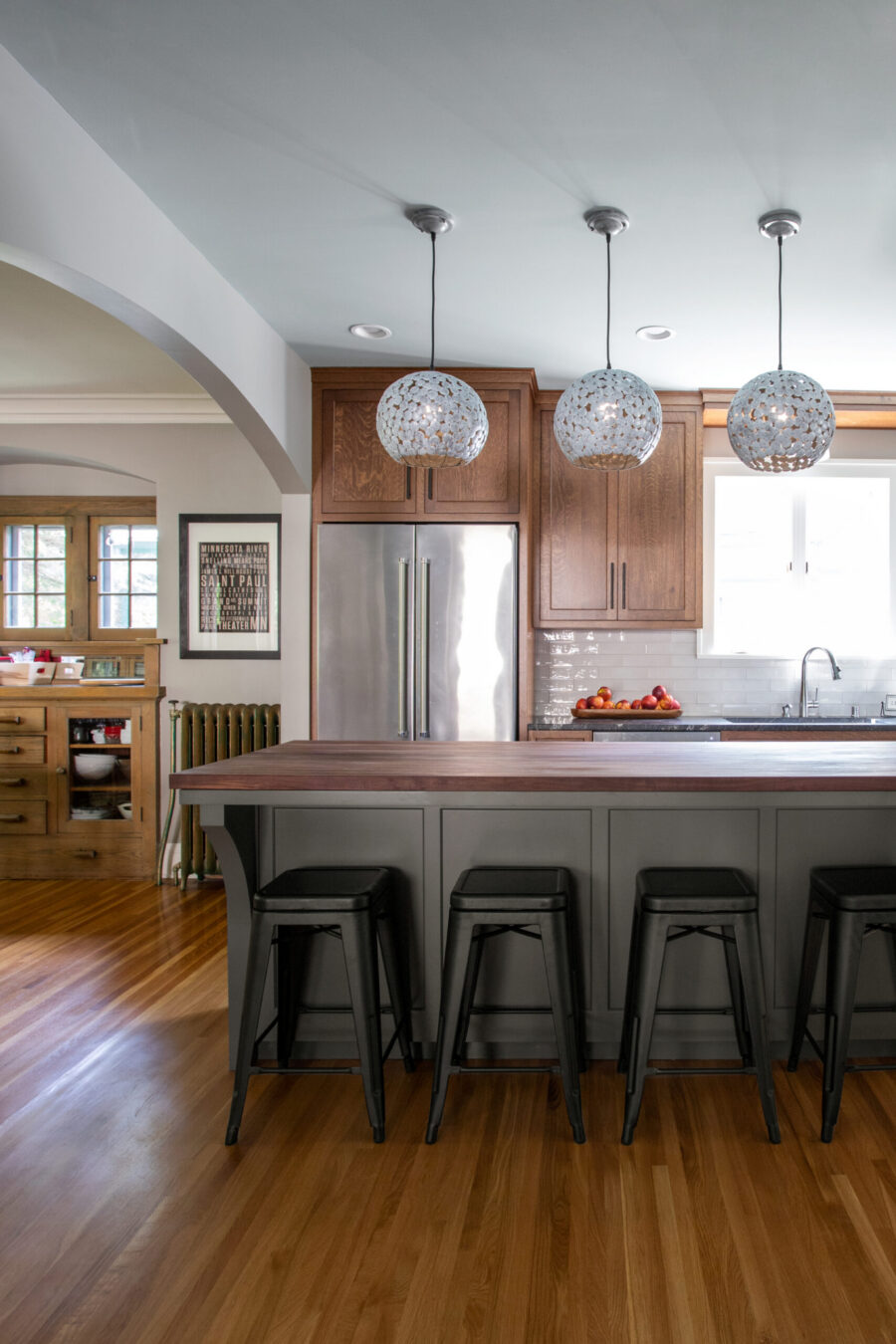 Twin Cities kitchen remodel. Wood cabinets, dark island with black stools. Three spherical pendant lights brighten white subway tile.